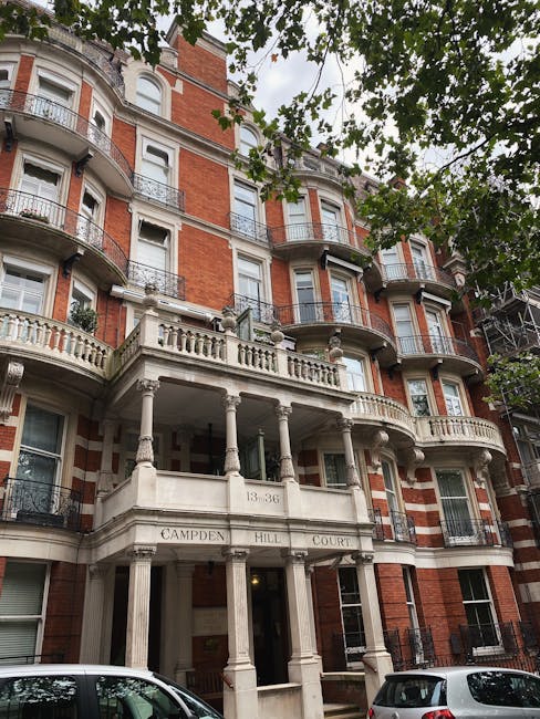  Exterior view of a multi-story red brick residential building on Campden Hill Court with ornate white stone balconies, columns, and decorative balustrades. The building features large sash windows with white frames, some of which have black wrought-iron railings. The facade shows signs of street-level accessibility, with a grand entrance supported by classical columns and the address number 33-36 displayed above. Green foliage from trees partially frames the scene, and parked cars are visible in the foreground. The image captures the architecture and clean, well-maintained appearance of the building, which may require specialized access solutions for flat cleaning or maintenance, as handled by Cleaner West Kensington.