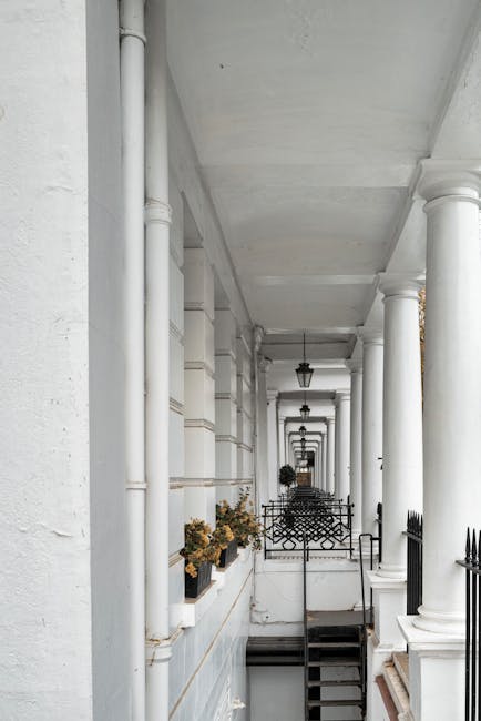 A white-painted exterior corridor featuring tall, round columns supporting an overhanging ceiling. The corridor has a row of potted plants with dried flowers placed on a ledge along the left side, and black metal fences with ornamental patterns on both sides. The surface appears clean and well-maintained, with natural daylight illuminating the space. The image presents a neat and tidy residential or commercial building facade, emphasizing surface cleanliness and structural detail, ideal for promoting services like surface cleaning or property maintenance by Cleaner West Kensington.