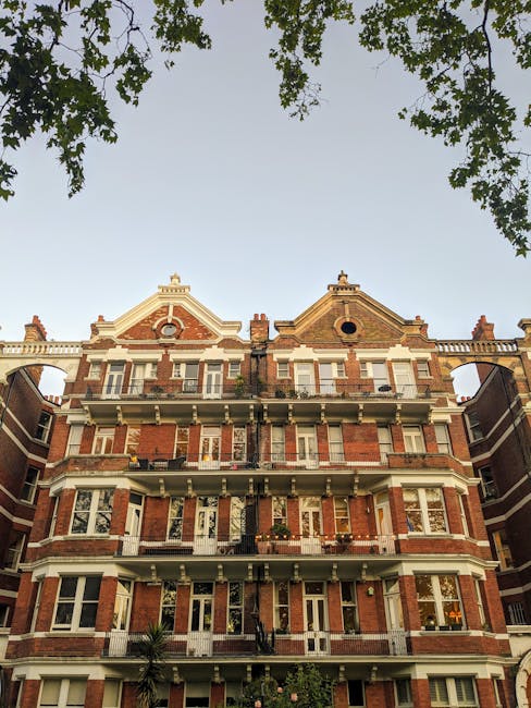 External view of a historic red brick residential building in Earls Court Kensington estates, displaying multiple floors with white decorative details, bay windows, balconies, and chimneys. The building is illuminated by natural daylight, with green tree branches visible at the top of the image. Cleaner West Kensington specializes in surface cleaning and deep cleaning services, maintaining the hygiene and appearance of such properties, although this image only depicts the building exterior. The overall scene showcases a well-maintained, classic London apartment block with clear, clean brickwork and tidy window areas.