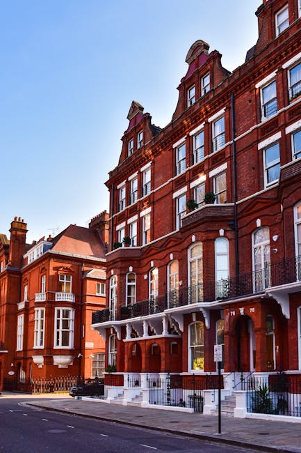 Exterior view of a red brick residential building on Kensington High Street showcasing multiple large windows, some with small decorative balconies with black wrought iron railings, and a prominent corner turret with a conical roof. The building features white trim accents around the windows and along the roofline, with a clean and well-maintained façade. The street in front is paved with asphalt, and there's a sidewalk with a parking restriction sign. Bright daylight and clear blue sky illuminate the scene, emphasizing the building's Victorian architectural details. The image highlights the historical charm and pristine condition of the building, in line with professional cleaning and maintenance standards promoted by Cleaner West Kensington for domestic and commercial properties.