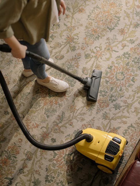 A person using a vacuum cleaner to perform deep cleaning on a patterned area rug with floral motifs in a residential living room. The individual is dressed in casual clothing, wearing white sneakers and beige trousers, and is holding the vacuum's handle while moving it across the beige and pastel-colored surface. The vacuum cleaner is yellow with a black hose, positioned on a wooden floor adjacent to the rug. The room is well-lit, showcasing a clean, freshly maintained space, with visible edges of furniture and a window reflecting natural light. This scene exemplifies professional surface cleaning by Cleaner West Kensington, emphasizing hygiene and meticulous stain removal on domestic carpets.
