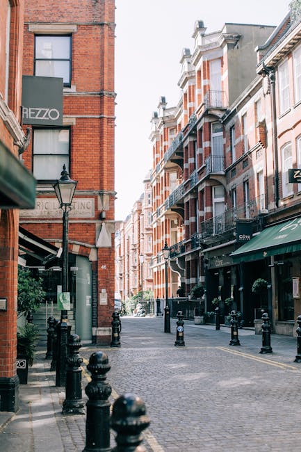 A quiet cobblestone street in WEST KENSINGTON lined with multi-story red brick buildings featuring white accents and black wrought iron balconies. The street is illuminated by natural daylight, with a