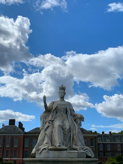 A beautifully detailed statue of Queen Victoria, made from light-colored stone, sits outdoors against a partly cloudy blue sky. The statue depicts the monarch seated, wearing a crown and royal robes, 
