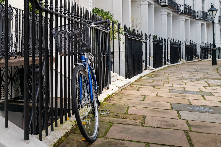 A residential street scene featuring a black wrought iron fence along a stone-paved sidewalk. A blue bicycle with a black wicker basket attached to the handlebars is parked against the fence near a white building. The pavement appears clean and well-maintained, with some moss or grass growing between the stone slabs. The building has white columns and window frames, with traditional black outdoor lamps mounted on the wall. Natural daylight illuminates the scene, highlighting the pristine condition of the surfaces. The image depicts an environment suitable for professional domestic or commercial surface cleaning and maintenance by Cleaner West Kensington.