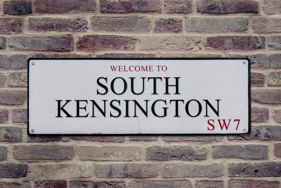 A rectangular street sign mounted on a brick wall, displaying the text 'Welcome to South Kensington' in black capital letters, with 'South Kensington' being prominently larger. The sign has a white background with red accents, including 'Welcome to' at the top and 'SW7' at the bottom right. The brick wall behind the sign features a pattern of irregularly textured, reddish-brown bricks, some with darker patches, giving a rustic appearance under natural lighting conditions.