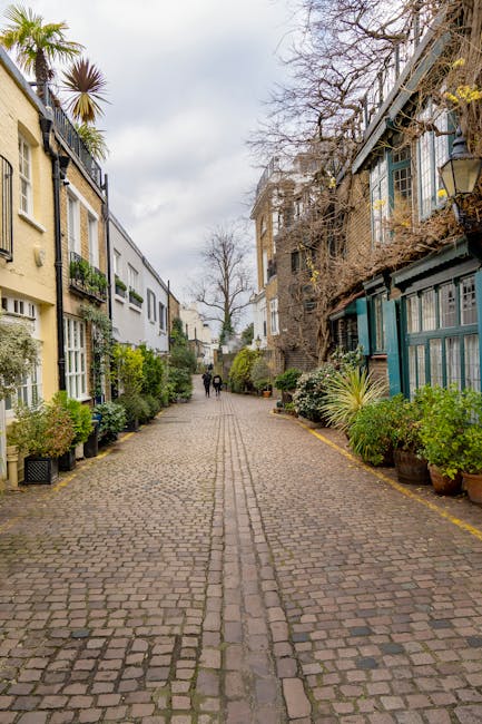 A cobblestone street in a residential area of Kensington, featuring a central pathway flanked by multi-story buildings with a mix of modern and traditional architecture. On the left, cream-colored bui