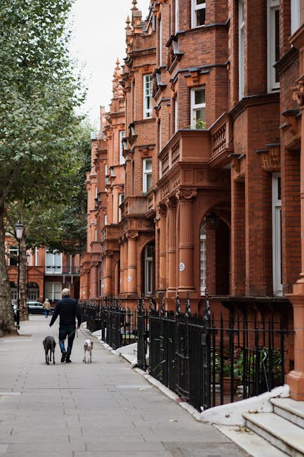 A cobblestone street in a residential area of Kensington, featuring a central pathway flanked by multi-story buildings with a mix of modern and traditional architecture. On the left, cream-colored bui