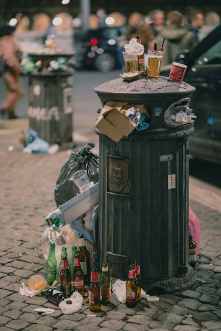 A pile of mixed household waste and recyclables scattered on a floor, including crumpled paper, plastic bottles, cardboard packaging, and used tissues. The surfaces appear cluttered and dirty, with so